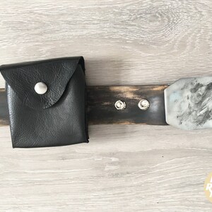 May include: A black leather belt with a silver buckle and a small black leather pouch. The belt is made of distressed wood and has two silver rivets. The pouch has a silver button.