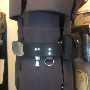 May include: A black leather belt with a silver buckle and two silver loops. The belt is worn on a mannequin and has a small black pouch attached to it.
