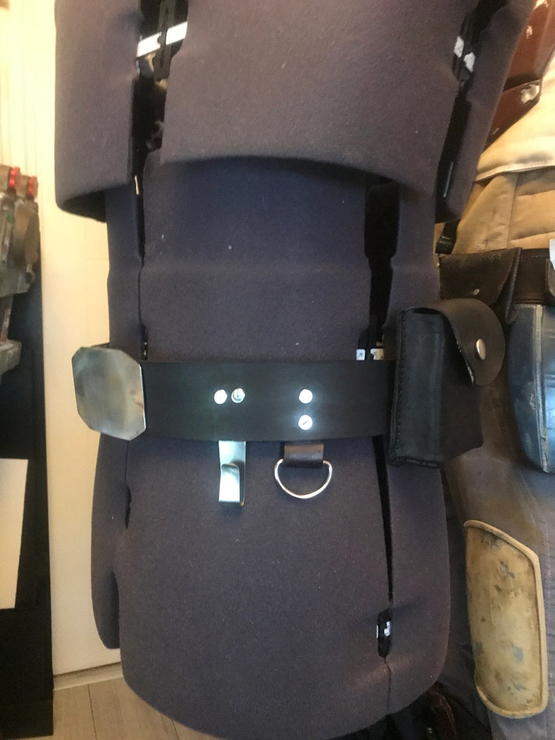 May include: A black leather belt with a silver buckle and two silver loops. The belt is worn on a mannequin and has a small black pouch attached to it.