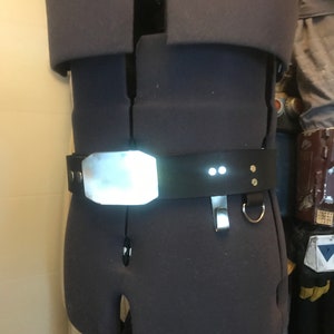 May include: A black leather belt with a silver buckle and two small white lights. The belt is worn on a mannequin.