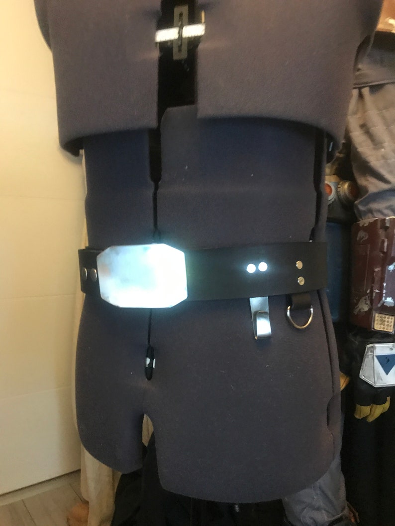 May include: A black leather belt with a silver buckle and two small white lights. The belt is worn on a mannequin.