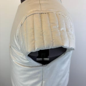 May include: Cream-colored garment with a padded shoulder detail. The shoulder area features vertical stitching, and the fabric has a subtle sheen. The garment is displayed on a mannequin, showcasing its shape and construction.