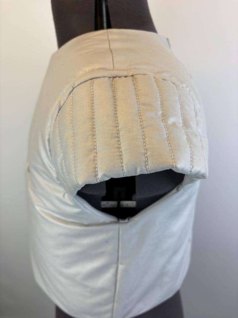 May include: Cream-colored garment with a padded shoulder detail. The shoulder area features vertical stitching, and the fabric has a subtle sheen. The garment is displayed on a mannequin, showcasing its shape and construction.