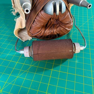 May include: A brown leather mask featuring a central circular opening with pleated leather edges, two metallic silver conical protrusions, and a cylindrical brown leather filter component attached by thin metal wires to the base of the mask.