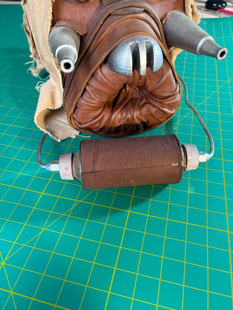 May include: A brown leather mask featuring a central circular opening with pleated leather edges, two metallic silver conical protrusions, and a cylindrical brown leather filter component attached by thin metal wires to the base of the mask.