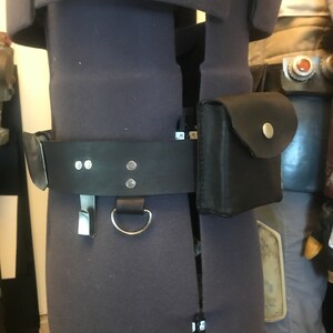 May include: A black leather belt with a silver buckle and a small black leather pouch attached to it. The belt is worn on a gray mannequin.