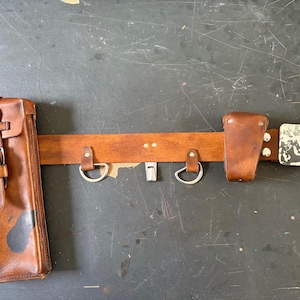 May include: A brown leather tool belt with two pouches and a metal buckle. The belt has two metal loops and a metal whistle holder.