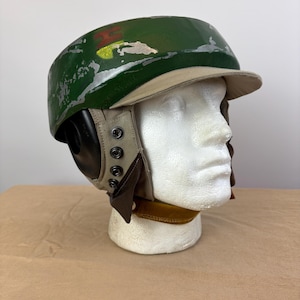 Puede incluir: Un casco verde con visera beige y orejeras. El casco tiene un acabado envejecido con pintura plateada y un diseño rojo, amarillo y verde en la parte superior. Las orejeras son negras y beige, y el casco se exhibe en una cabeza de maniquí blanca.