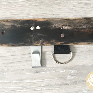 May include: A black wooden shelf with two metal hooks and a leather strap with a metal ring. The shelf has a distressed finish and is mounted on a light wood wall.