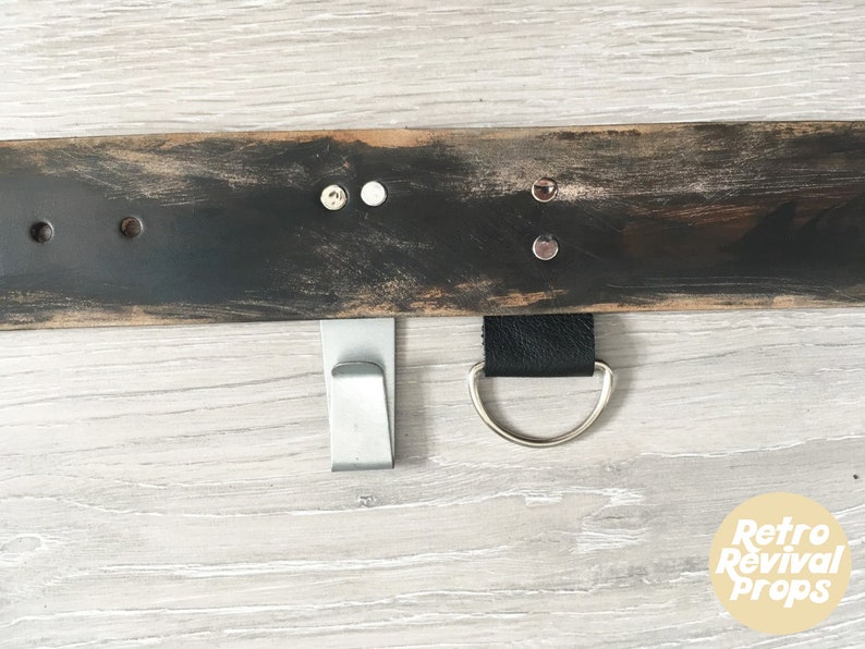 May include: A black wooden shelf with two metal hooks and a leather strap with a metal ring. The shelf has a distressed finish and is mounted on a light wood wall.