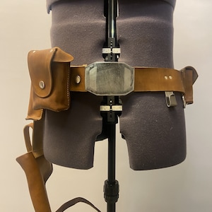 May include: A brown leather belt with a silver buckle and a brown leather holster attached to the left side. The belt is worn on a gray mannequin.