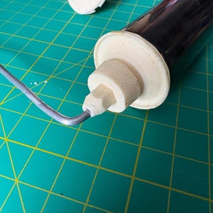 May include: A kitchen tool featuring a dark brown cylindrical body with off-white plastic end caps and a threaded nozzle. A thin, bent silver-colored metal wire frame is attached to the side of the nozzle, extending outward from the main body.