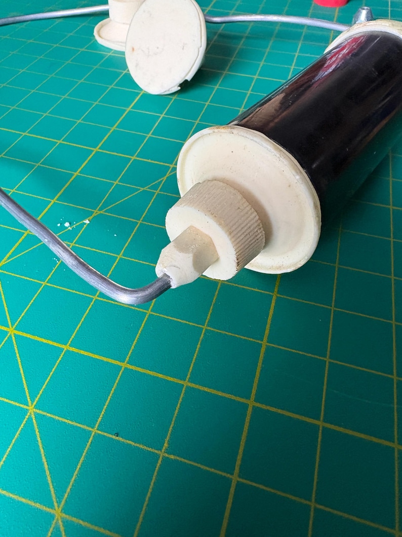 May include: A kitchen tool featuring a dark brown cylindrical body with off-white plastic end caps and a threaded nozzle. A thin, bent silver-colored metal wire frame is attached to the side of the nozzle, extending outward from the main body.