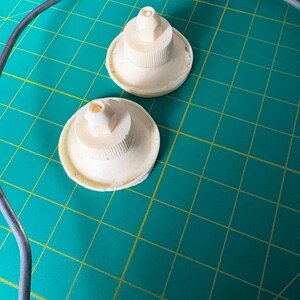 May include: Two off-white, circular plastic components with ribbed, cylindrical centers and small, pointed nozzles at the top. These items are positioned near a curved, thin, grey metal wire frame, showcasing a functional design.