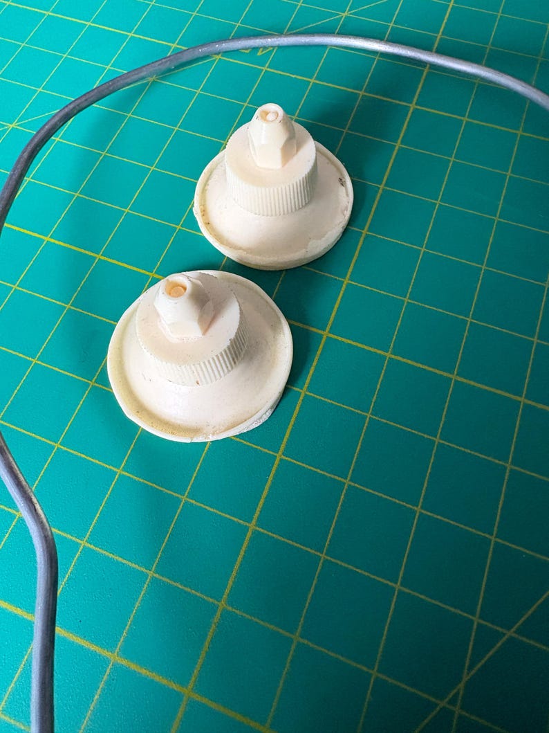 May include: Two off-white, circular plastic components with ribbed, cylindrical centers and small, pointed nozzles at the top. These items are positioned near a curved, thin, grey metal wire frame, showcasing a functional design.