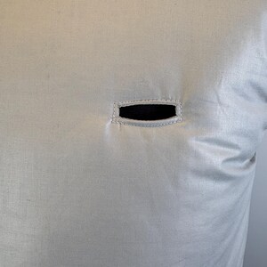 May include: Close-up of a light beige fabric with a rectangular black opening. The opening is framed by a stitched border. The fabric has a subtle sheen and texture, suggesting a pillow or cushion cover.