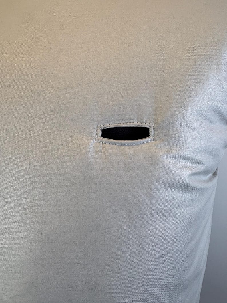 May include: Close-up of a light beige fabric with a rectangular black opening. The opening is framed by a stitched border. The fabric has a subtle sheen and texture, suggesting a pillow or cushion cover.