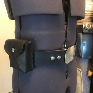 May include: A black leather belt with a silver buckle and two silver studs. A black leather pouch with a flap and a silver button is attached to the belt.