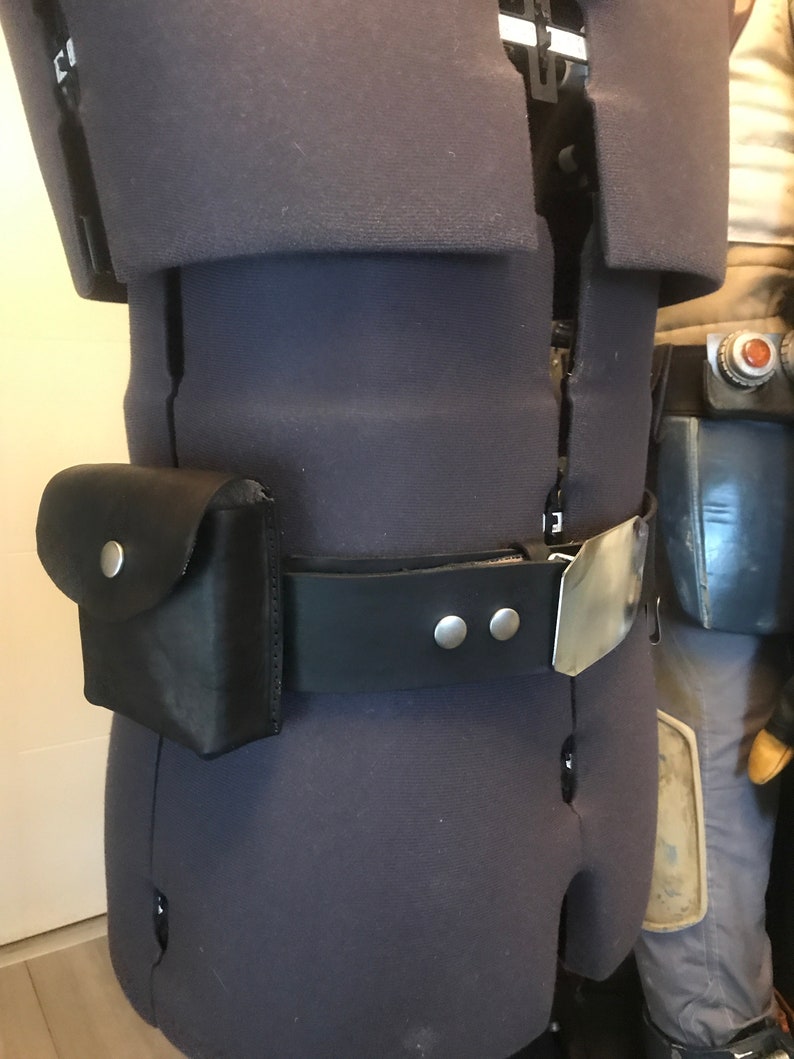 May include: A black leather belt with a silver buckle and two silver studs. A black leather pouch with a flap and a silver button is attached to the belt.
