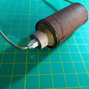 May include: A cylindrical object wrapped in a textured brown material, featuring a metallic end cap with a ribbed edge and a silver-toned nozzle. A thin, dark, curved wire extends from the center of the nozzle, creating a loop that arches outward.