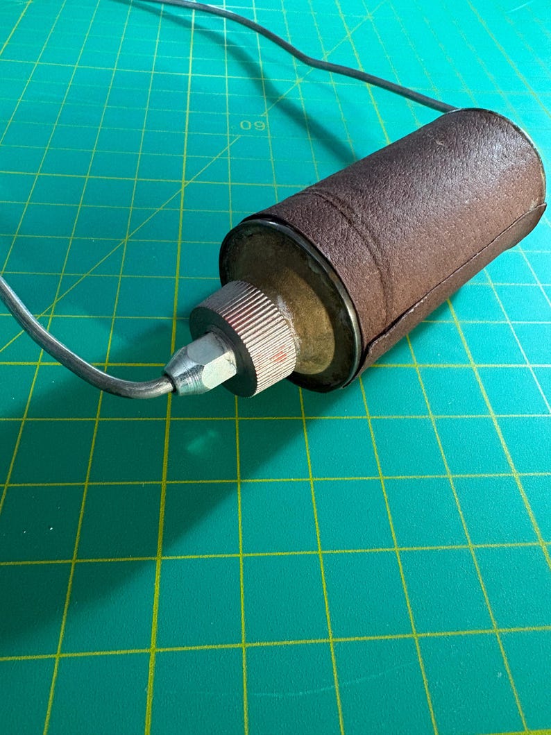 May include: A cylindrical object wrapped in a textured brown material, featuring a metallic end cap with a ribbed edge and a silver-toned nozzle. A thin, dark, curved wire extends from the center of the nozzle, creating a loop that arches outward.
