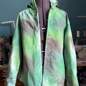 May include: A green and gray tie-dye hooded jacket displayed on a mannequin. The jacket has a drawstring hood, long sleeves, and a button-up front. The fabric has a gradient effect, transitioning from green to gray.
