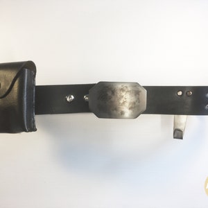 May include: A black leather belt with a silver buckle and a small black leather pouch attached. The belt has silver rivets and a silver hook.