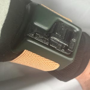 May include: A dark green wrist-worn device with a textured tan strap. The device has a rectangular shape with a detailed black design on its surface. The wristband is secured on a black and white striped sleeve.