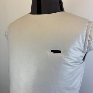 May include: A light gray, quilted vest with a small, horizontal opening. The vest is displayed on a black dress form. The vest has a rounded neckline and short sleeves. The fabric has a subtle sheen.