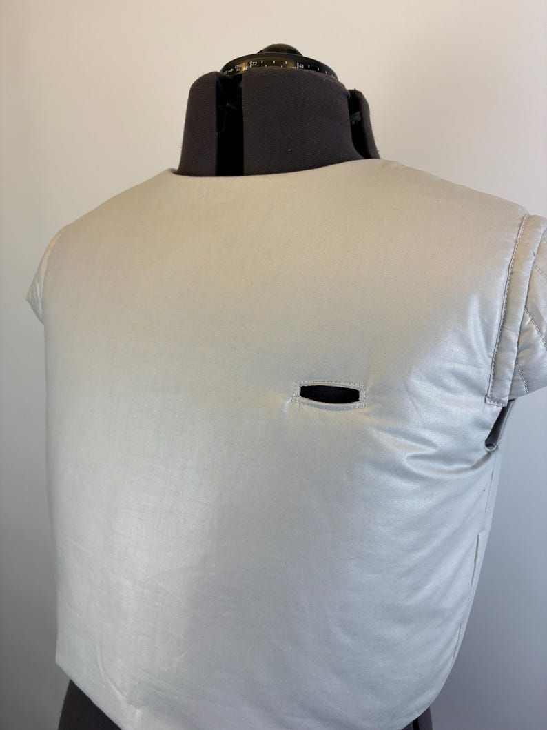 May include: A light gray, quilted vest with a small, horizontal opening. The vest is displayed on a black dress form. The vest has a rounded neckline and short sleeves. The fabric has a subtle sheen.