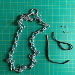 May include: A silver metal dog chain lead with a clasp and a handle, alongside two safety pins and a black leather strap, arranged on a green grid surface. The chain is made of interlocking links.