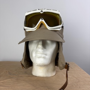 May include: A vintage aviator hat with attached goggles. The hat is a muted olive green with ear flaps and a front brim. The goggles have a white frame and yellow-tinted lenses. A brown leather strap with a metal clasp is attached to the hat.