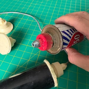 May include: A metal canister with a red cap and a silver nozzle, featuring blue and white diagonal stripes and the partial text OSS. Two additional white plastic nozzles and a black cylindrical tool are present, along with a brown clay-like object.