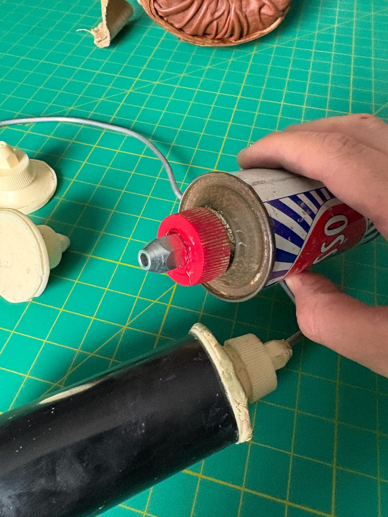 May include: A metal canister with a red cap and a silver nozzle, featuring blue and white diagonal stripes and the partial text OSS. Two additional white plastic nozzles and a black cylindrical tool are present, along with a brown clay-like object.
