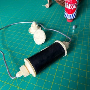 May include: A black cylindrical component with cream-colored plastic ends attached to a thin silver metal wire frame. Nearby are two cream-colored plastic circular pieces, a small silver metal nut, and a metal can labeled Brasso with red and blue.