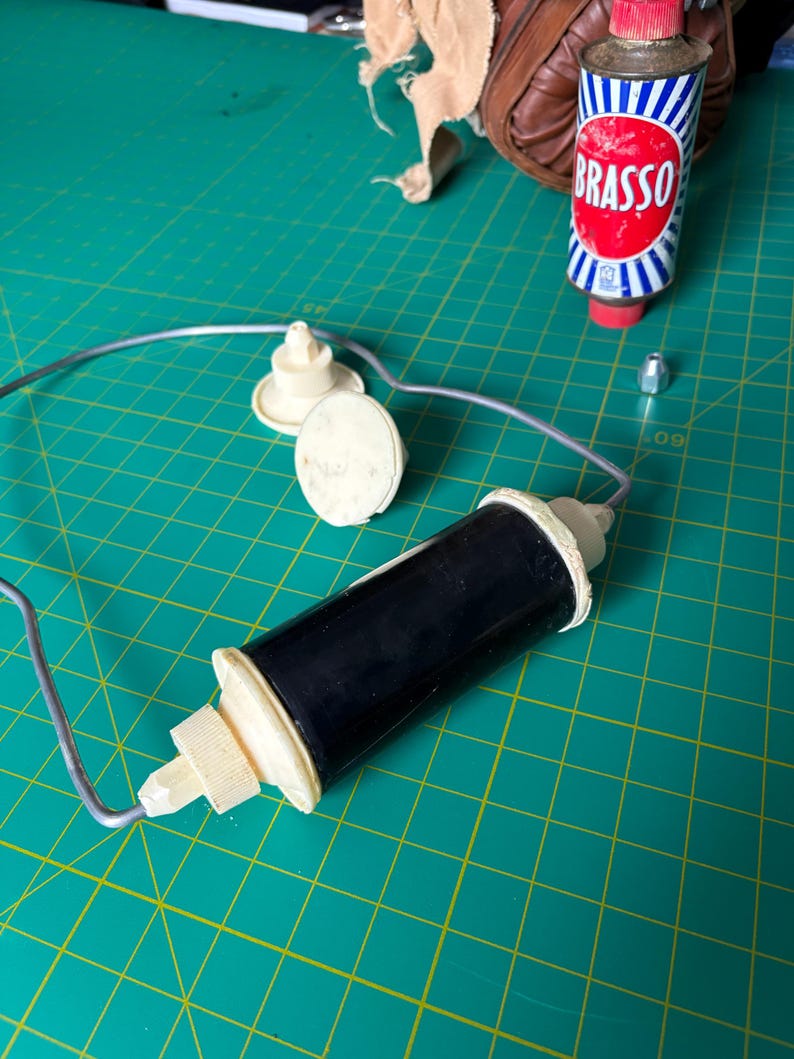 May include: A black cylindrical component with cream-colored plastic ends attached to a thin silver metal wire frame. Nearby are two cream-colored plastic circular pieces, a small silver metal nut, and a metal can labeled Brasso with red and blue.