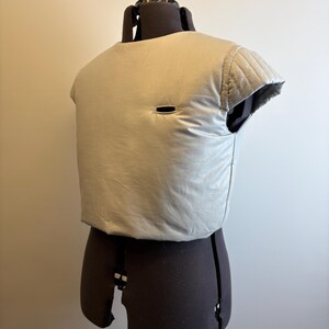 May include: A light gray, padded vest with a small rectangular opening. The vest is displayed on a dark brown dress form. The vest has quilted detailing on the shoulders and a slightly rounded neckline. The fabric appears to be a soft, matte material.