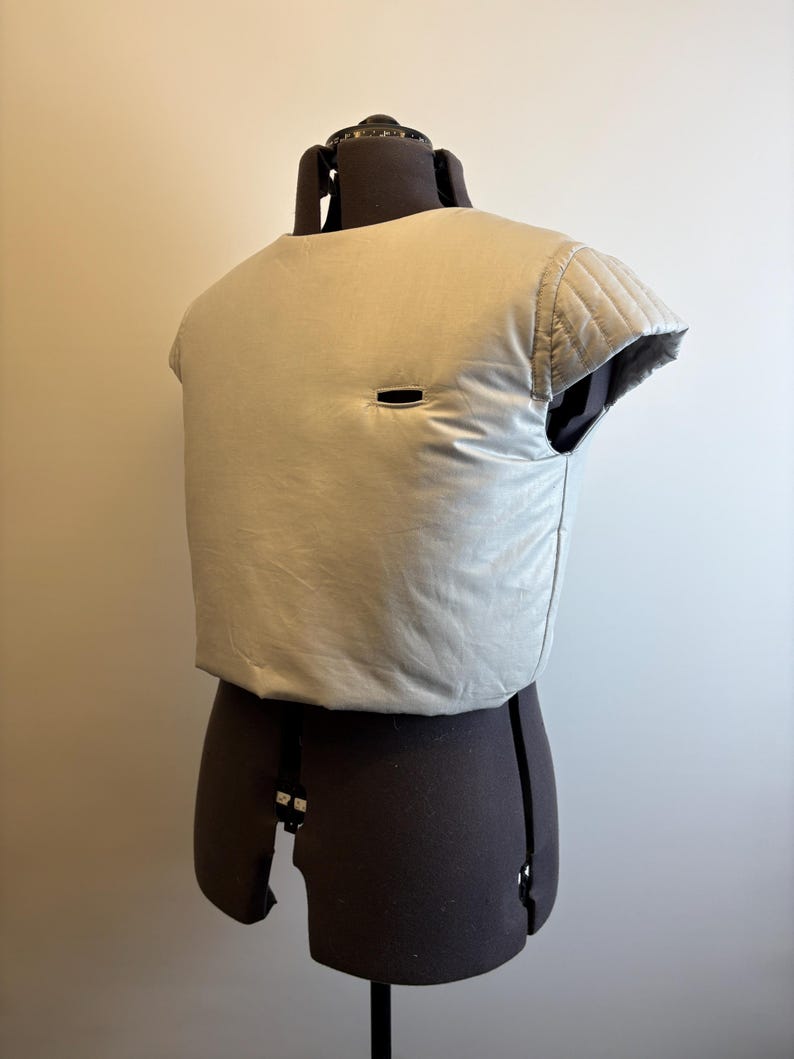May include: A light gray, padded vest with a small rectangular opening. The vest is displayed on a dark brown dress form. The vest has quilted detailing on the shoulders and a slightly rounded neckline. The fabric appears to be a soft, matte material.