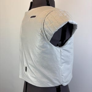 May include: A light silver vest with a quilted shoulder detail and a small rectangular opening. The vest is displayed on a black dress form against a plain white background. The vest has a shiny, smooth texture.