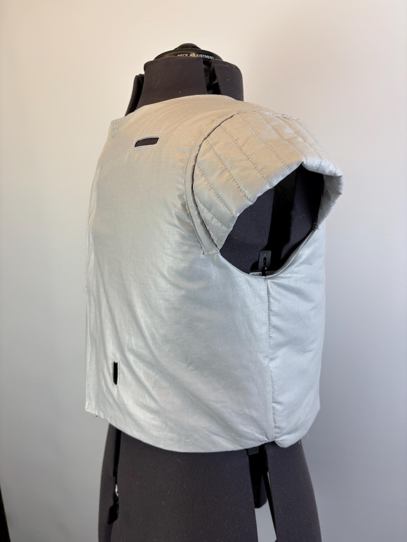 May include: A light silver vest with a quilted shoulder detail and a small rectangular opening. The vest is displayed on a black dress form against a plain white background. The vest has a shiny, smooth texture.
