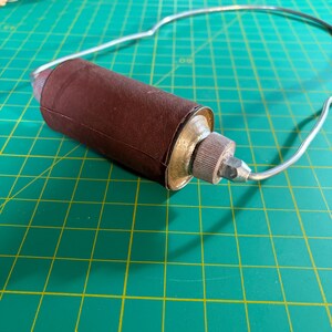 May include: A handheld sanding tool featuring a cylindrical brown abrasive sleeve wrapped around a central core, attached to a curved silver metal wire frame with a textured metallic knob and a small pointed tip at the end of the assembly.