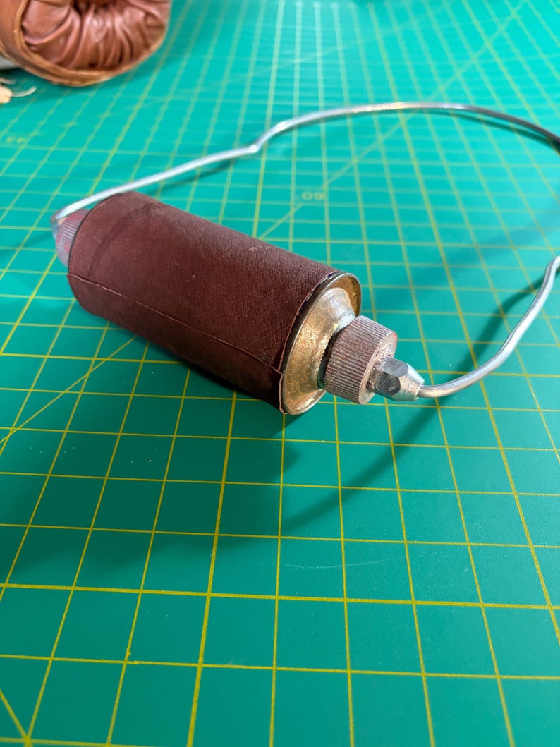 May include: A handheld sanding tool featuring a cylindrical brown abrasive sleeve wrapped around a central core, attached to a curved silver metal wire frame with a textured metallic knob and a small pointed tip at the end of the assembly.