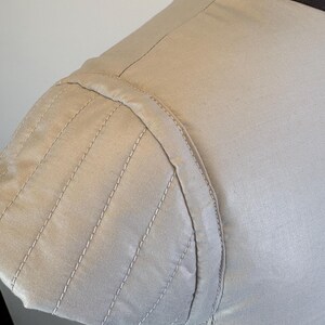 May include: Close-up of a beige garment's shoulder and sleeve. The sleeve features a series of parallel, stitched lines, creating a textured effect. The stitching is done in a lighter shade, providing contrast. The fabric appears to be a smooth, woven material.