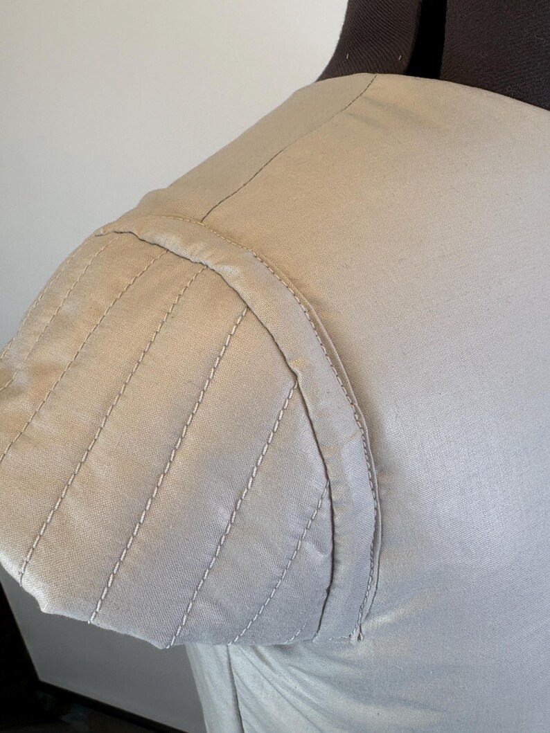 May include: Close-up of a beige garment's shoulder and sleeve. The sleeve features a series of parallel, stitched lines, creating a textured effect. The stitching is done in a lighter shade, providing contrast. The fabric appears to be a smooth, woven material.