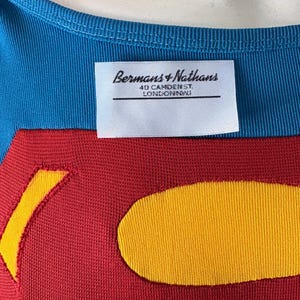 May include: Close-up of a blue and red garment with a yellow emblem. The garment has a blue neckline and a red body with a yellow emblem. A white label reads "Bermans + Nathans 40 Camden St. LONDON NW1".