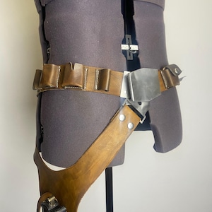 May include: A brown leather belt with a metal buckle and a holster for a weapon. The belt is worn on a mannequin.