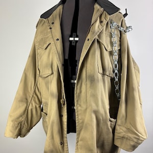 May include: A tan military-style jacket with a black collar and a silver chain detail. The jacket has multiple pockets, a zipper closure, and a drawstring at the bottom. The jacket is displayed on a black mannequin.