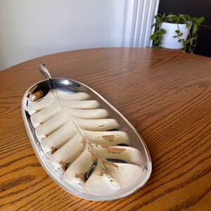 May include: A silver leaf-shaped serving tray with a raised design. The tray is oval and has a handle on one end.