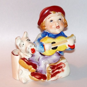 May include: A ceramic figurine of a child wearing a red hat and playing a yellow guitar, sitting next to a white dog. The figurine is a vintage style and has a hand-painted look.