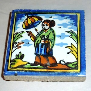 May include: A square ceramic tile with a blue border and a colorful illustration of a person holding an umbrella. The person is wearing a green and blue robe and has brown hair. The background is a blue sky with white clouds and green plants.
